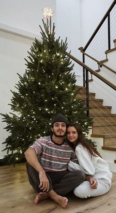 a man and woman sitting in front of a christmas tree on the floor next to stairs