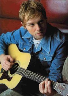 a man sitting on a couch holding a guitar in his hand and looking at the camera