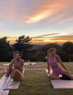 two women sitting on yoga mats in the middle of a field at sunset, one talking on her cell phone