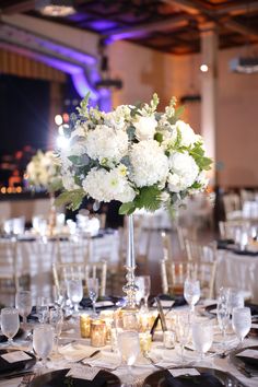 white flowers are in a tall glass vase on the center table at a wedding reception