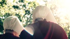 two women dressed in white are walking through the sunlit forest with their backs to each other