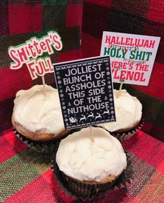 three cupcakes with white frosting and small signs on them sitting on a plaid table cloth