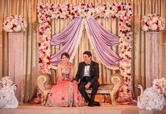 a man and woman sitting on a bench in front of a floral decorated stage with flowers