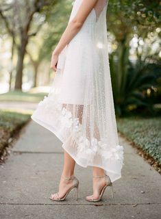 a woman wearing high heels and a white dress with flowers on the bottom, standing in front of trees