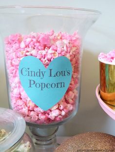 pink and yellow popcorn in a glass bowl with a blue heart shaped sign on it