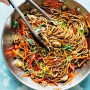 two hands holding chopsticks over a bowl of noodles and vegetables with mushrooms on the side