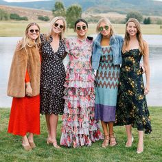 four women standing next to each other in front of a lake wearing dresses and fur coats