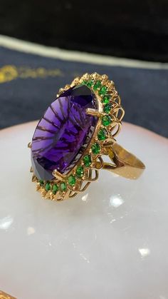 An exquisite piece to make a statement anywhere you go. Whether your style is a brooch, pendant, or ring, it can do it all. 

💚 Stone description:

Centre stone: Natural Amethyst

Shape: Classic Oval cut

Weight: 59.26CTS

Dimensions: 28 X 21 MM

Clarity: Eye-Clean

Origin: Brazil

Treatment: None

Hardness: 8

Hue: Intense Purple

Quality: Top notch

🤍About side Stones:

Stone: Natural Tsavorite

Shape: Brilliant Round cut

Weight: 3.92cts

Quality: Extra fine

💛About Metal:

Metal: 18k Solid Yellow Gold

Ring size: 6 US Gold Rings With Stones, Beautiful Jewelry Unique, Amethyst Jewelry Ring, Yellow Stone Ring, Yellow Gold Amethyst Ring, Stone Ring Design, Tsavorite Ring, Amethyst Rings, Amethyst Cocktail Ring