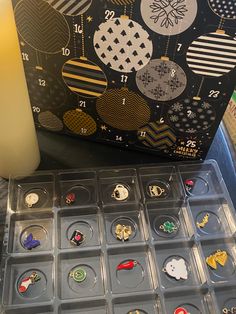 an assortment of christmas themed pins and buttons in a box next to a candle on a table