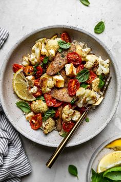 a white bowl filled with chicken, cauliflower and tomato salad next to lemon wedges