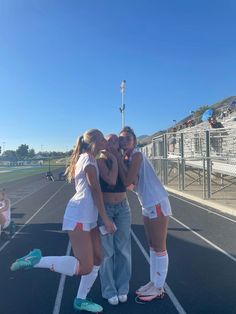 three girls are hugging on the side of a tennis court while one girl is wearing white shorts