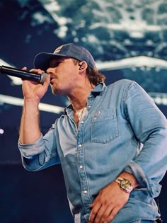 a man singing into a microphone while wearing a denim shirt and hat with his hands in the air