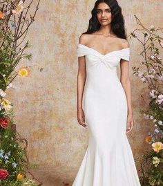 a woman standing in front of flowers wearing a white dress with off the shoulder sleeves