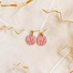 two pink earrings with leaves on them sitting on top of a white cloth next to streamers