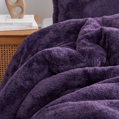 a purple comforter on a bed next to a wooden table with a book and lamp