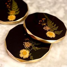 three black and gold coasters with yellow daisies on the front one has green leaves