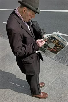 an old man in a suit and hat is reading a newspaper on the side walk