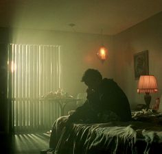 a man sitting on top of a bed next to a table with a lamp in it