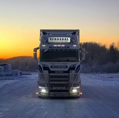 a semi truck driving down a snow covered road at sunset with the sun setting in the background