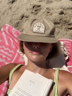 a woman laying in the sand with a book and hat on her head while reading