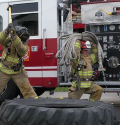 two firemen in front of a fire truck with their hoses on and one holding a large tire