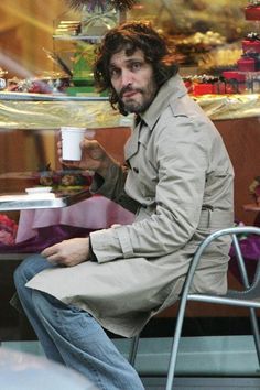 a man sitting at a table with a coffee cup in his hand and wearing a trench coat