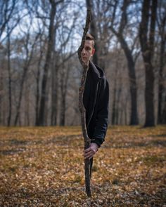 a young man holding onto a tree branch in the middle of a forest with leaves on the ground