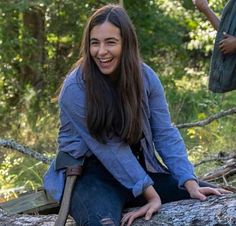 a woman sitting on top of a log in the woods smiling at someone behind her