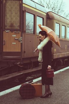 a woman holding an umbrella standing next to a train car with luggage on the ground