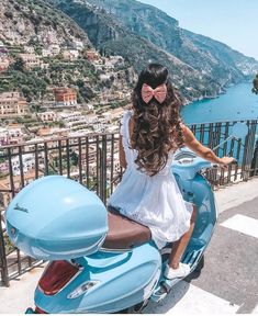 a woman sitting on top of a blue scooter next to the ocean and mountains