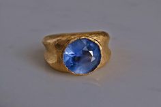 Fine 24k gold ring set with an unheated Sri Lankan sapphire weighing 7.5ct. The round blue sapphire is light cornflower blue. The stone has medium saturation, but its light cornflower hue is unique. This unique blue wrapped by the purity of gold sets up a ring with opposing lights. The stone is cool and the gold is warm - the ring carries a light stone, as well as a light-infused stone. In any hue of blue, a round sapphire is harder to find than one can imagine. The band is reminiscent of Roman rings, except an actual Roman ring will not be as hefty. The ring now weighs 21.69 grams, a size 7. This design is built on ideas of continuity, the round stone flowing to its sculpted gold work. At this stone's size, its design will take any finger size, it is quintessentially Zen. The stone measur Yellow Gold Sapphire Ring With Faceted Detail, Blue Faceted Sapphire Ring Fine Jewelry, Blue Faceted Sapphire Ring In Fine Jewelry Style, Handmade Yellow Gold Sapphire Ring, Formal Faceted Blue Sapphire Ring, Formal Blue Faceted Sapphire Ring, Faceted Yellow Gold Sapphire Ring, Blue Faceted Sapphire Ring For Wedding, Gold Sapphire Rings With Faceted Detail