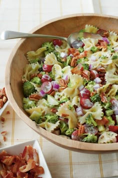 a wooden bowl filled with pasta salad next to a plate of bacon and broccoli