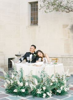 a bride and groom sitting at a table in front of a building with white flowers