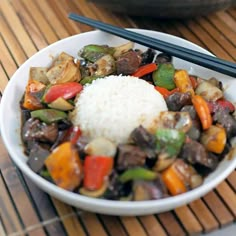 a white bowl filled with vegetables and rice next to chopsticks on a mat