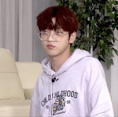 a young man with red hair and glasses sitting in front of a white couch wearing a hoodie