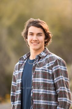 a young man standing in front of a forest wearing a flannel shirt and jeans