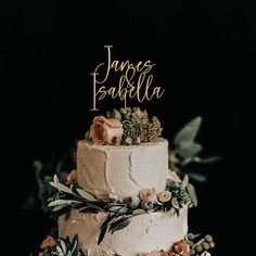 a three tiered wedding cake with flowers and greenery on the top is decorated with gold lettering