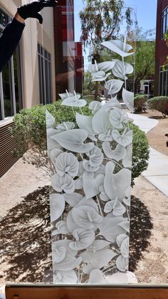 a glass sculpture with white flowers on it in front of a building and some bushes