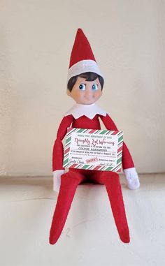 an elf is sitting on the shelf holding a sign