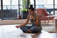 a woman is sitting on the floor looking at her cell phone and drinking from a cup