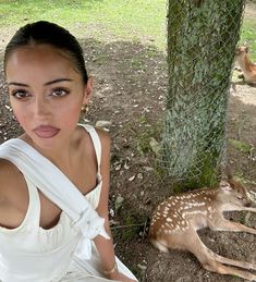 a woman in white dress sitting next to a tree with deer on the ground behind her