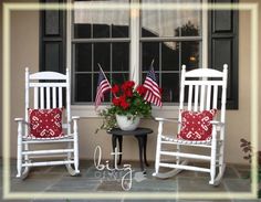 two white rocking chairs with red and white pillows sitting on a porch next to an american flag potted plant