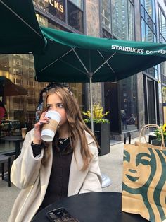 a woman drinking from a coffee cup while sitting at an outdoor table in front of starbucks