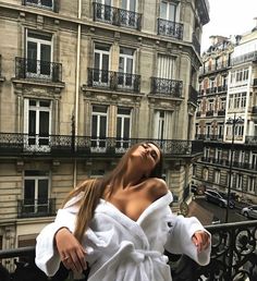 a woman in white robe standing on balcony next to building and looking up at the sky