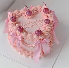 a heart shaped cake with pink frosting and cherries on top, sitting on a white surface