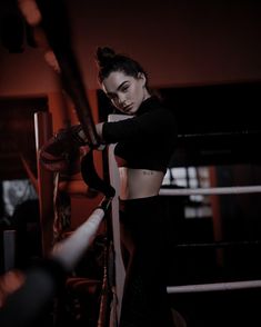 a woman in black top and leggings standing next to a boxing ring with her hand on the ropes