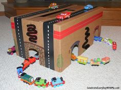 a cardboard box that has cars and trucks in it on the floor next to a toy train set