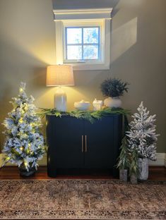 two christmas trees sitting on top of a rug next to a cabinet and lamp in a room