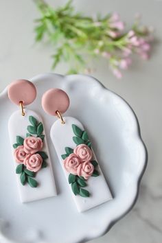 two white and pink earrings with roses on them sitting on a plate next to flowers