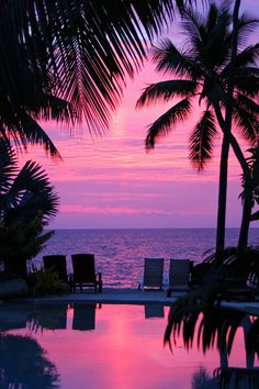 the sun is setting behind some palm trees and lounge chairs by the water's edge